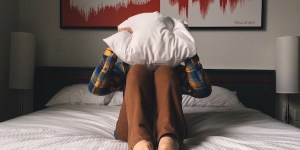 Frustrated man sitting on top of bed and hiding head under pillow