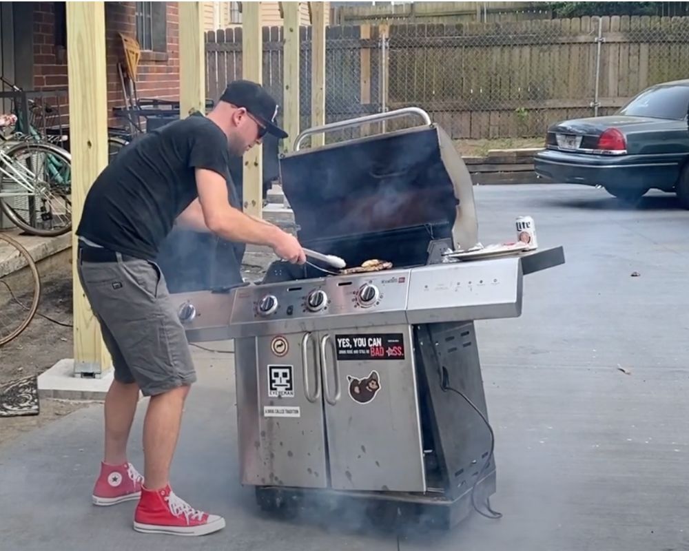 Man grilling on a broken propane grill that needs to be disposed of.